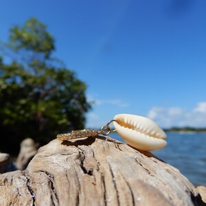Peut inclure: Un pendentif en or avec un petit charme de coquillage blanc. Le pendentif repose sur un morceau de bois flotté avec une étendue d'eau bleue en arrière-plan.