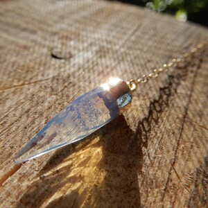 Peut inclure: Un pendule en cristal clair en forme de goutte d'eau, avec un capuchon et une chaîne dorés. Le cristal a une teinte bleu clair et est suspendu sur un fond en bois naturel, captant la lumière du soleil.