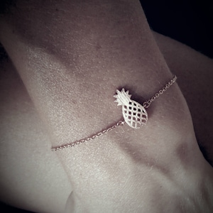 May include: A gold pineapple charm bracelet with a delicate chain. The bracelet is worn on a person's wrist.