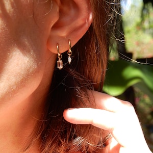 Peut inclure: Gros plan sur une oreille ornée de deux boucles d'oreilles créoles dorées. L'une comporte une pierre rectangulaire transparente, l'autre une petite gemme en forme de goutte. Les boucles d'oreilles sont délicates et élégantes.