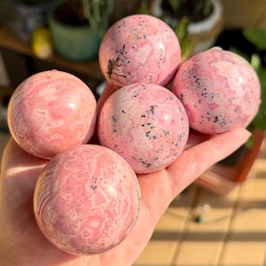 以下が含まれることがあります： 鮮やかなピンク色のパレットに渦巻く模様と黒い鉱物の内包物が散りばめられた、5つの磨かれた球状の石。それぞれの球体は滑らかで光沢のある表面の質感を持ち、ピンクと白の石の自然な変化を見せています。