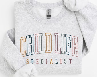 Może przedstawiać: Szara bluza z nadrukiem "CHILD LIFE SPECIALIST" w kolorowym, uniwersyteckim stylu.