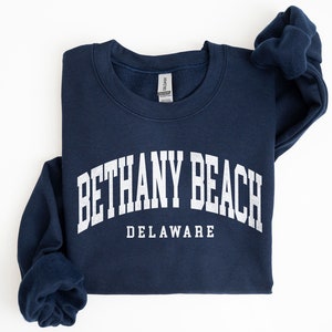 May include: A navy blue sweatshirt with the text "BETHANY BEACH DELAWARE" in white, arched across the front. The sweatshirt is folded, with the sleeves and collar visible against a white background.