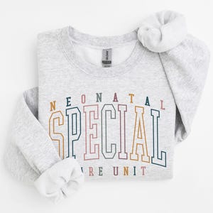 May include: A light gray sweatshirt with the words "NEONATAL SPECIAL CARE UNIT" in a colorful, collegiate-style font. The sweatshirt has a crew neck and long sleeves with rolled cuffs. The Gildan brand tag is visible.