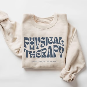 May include: A beige sweatshirt with the text "PHYSICAL THERAPY" in a blue retro font. The text is followed by "GROSS MOTOR PROMOTER" in smaller blue text.