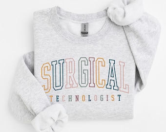 May include: A light gray crewneck sweatshirt featuring the text SURGICAL TECHNOLOGIST in a collegiate-style font. The word SURGICAL is arched in large, multicolored block letters, while TECHNOLOGIST appears below in smaller, solid-colored letters.