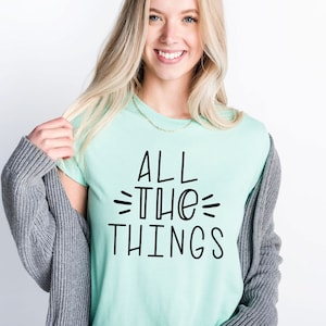 May include: A woman wearing a light green t-shirt with the text "ALL THE THINGS" in black lettering. She is smiling and has blonde hair. She is wearing a gray cardigan over the t-shirt.