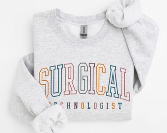 May include: A light gray crewneck sweatshirt featuring the text SURGICAL TECHNOLOGIST printed in a collegiate font. The letters are outlined in various colors including dark blue, orange, and muted red. The garment has long sleeves.