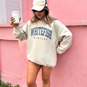 May include: Cream-colored sweatshirt with the text "WHITEFISH MONTANA" in navy blue. The person in the image is wearing a white baseball cap with a black "A" on it and sunglasses. They are holding a white coffee cup.