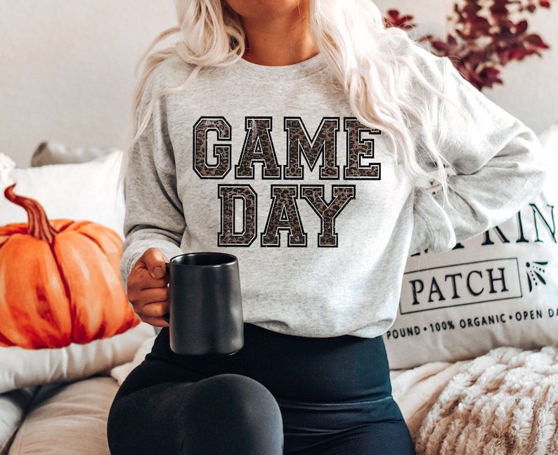 Pu&ograve; includere: Una felpa grigia con la scritta "GAME DAY" stampata in nero con un motivo leopardato.