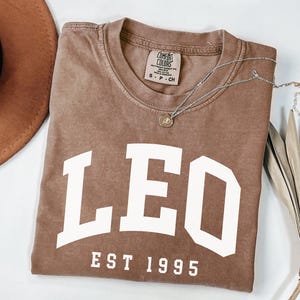 May include: A brown Comfort Colors t-shirt with the word "LEO" in large white letters and "EST 1995" below. A silver necklace with a small pendant rests on the shirt. A brown hat and dried palm leaf are in the background.