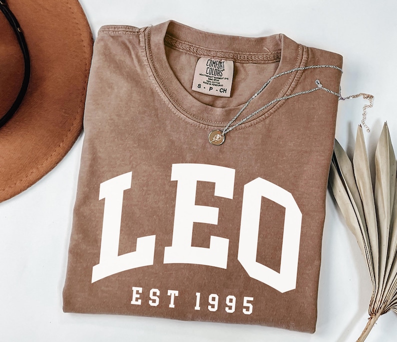 May include: A brown Comfort Colors t-shirt with the word "LEO" in large white letters and "EST 1995" below. A silver necklace with a small pendant rests on the shirt. A brown hat and dried palm leaf are in the background.