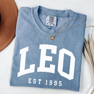 May include: A blue Comfort Colors t-shirt with the word "LEO" in large white letters, and "EST 1995" below. A gold necklace with a pendant rests on the shirt. A brown hat and dried plant are in the background.