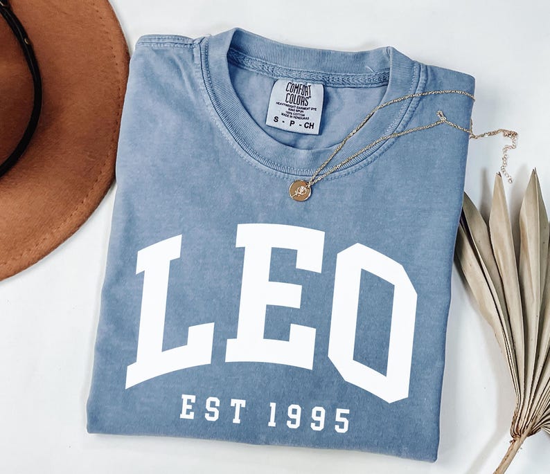 May include: A blue Comfort Colors t-shirt with the word "LEO" in large white letters, and "EST 1995" below. A gold necklace with a pendant rests on the shirt. A brown hat and dried plant are in the background.