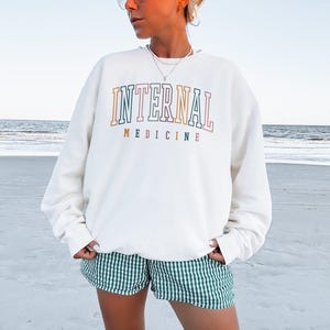 Puede incluir: Sudadera de color crema con las palabras "INTERNAL MEDICINE" en letras coloridas. La persona lleva unos pantalones cortos a cuadros verdes y blancos. El fondo es una playa con el oc&eacute;ano al fondo.