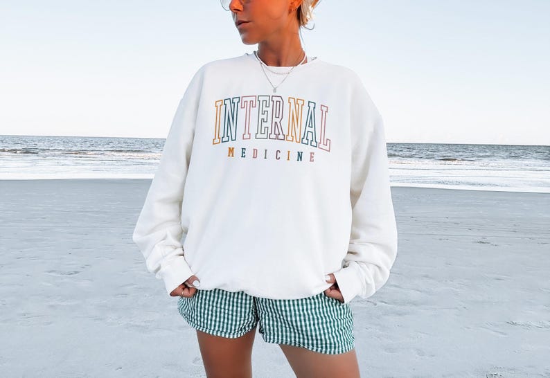 Puede incluir: Sudadera de color crema con las palabras "INTERNAL MEDICINE" en letras coloridas. La persona lleva unos pantalones cortos a cuadros verdes y blancos. El fondo es una playa con el oc&eacute;ano al fondo.