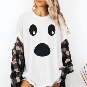 May include: White t-shirt featuring a surprised ghost face design with large black oval eyes and a large black oval mouth. The shirt is a Halloween-themed graphic tee.