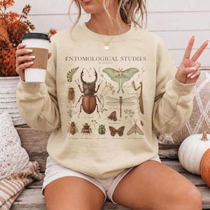 May include: A beige crewneck sweatshirt featuring a graphic print titled ENTOMOLOGICAL STUDIES PLATE XLVII. The design displays various detailed illustrations of insects, including a large stag beetle, a moth, a dragonfly, and a praying mantis.