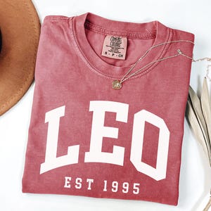 May include: A dusty rose-colored t-shirt with the word "LEO" in large white block letters. Below the word "LEO" is the text "EST 1995". A gold necklace with a pendant is draped across the shirt. A brown hat and dried leaves are in the background.
