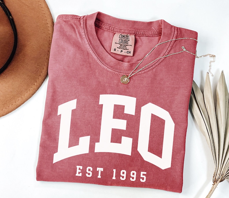 May include: A dusty rose-colored t-shirt with the word "LEO" in large white block letters. Below the word "LEO" is the text "EST 1995". A gold necklace with a pendant is draped across the shirt. A brown hat and dried leaves are in the background.