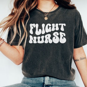 May include: A dark gray t-shirt with the text "FLIGHT NURSE" in white, retro-style lettering. The shirt is worn by a person with long, wavy hair.