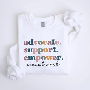 May include: White sweatshirt with the words "advocate. support. empower. social work" in a colorful, stacked design. The text is in various colors, including teal, pink, and gold. The sweatshirt has long sleeves and a crew neck.