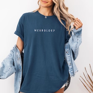 K&ouml;nnte beinhalten: Eine Frau tr&auml;gt ein dunkelblaues T-Shirt mit dem Wort "NEUROLOGY" in wei&szlig; auf der Vorderseite. Sie tr&auml;gt eine hellblaue Jeansjacke &uuml;ber dem Shirt.