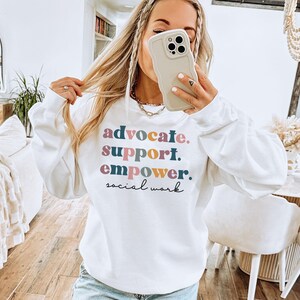 May include: White sweatshirt with the words "advocate. support. empower. social work" printed in colorful text. The sweatshirt has long sleeves and a crew neck.