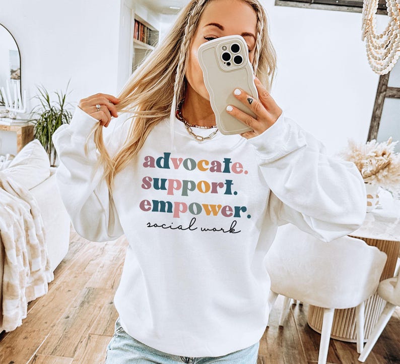 May include: White sweatshirt with the words "advocate. support. empower. social work" printed in colorful text. The sweatshirt has long sleeves and a crew neck.