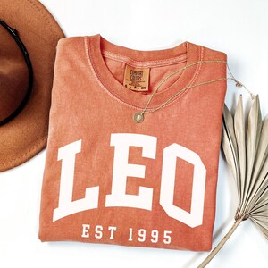 May include: A coral-colored t-shirt with the word "LEO" in large white block letters and "EST 1995" underneath. A gold necklace with a pendant and a brown hat are next to the shirt. The shirt has a Comfort Colors tag.