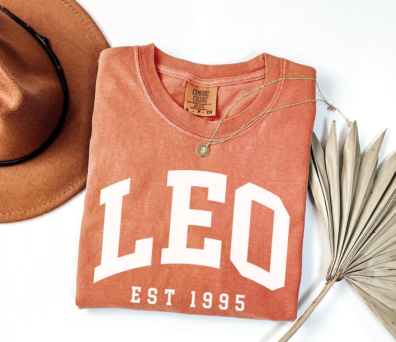 May include: A coral-colored t-shirt with the word "LEO" in large white block letters and "EST 1995" underneath. A gold necklace with a pendant and a brown hat are next to the shirt. The shirt has a Comfort Colors tag.
