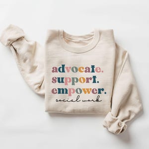 May include: Cream-colored sweatshirt with the words "advocate. support. empower. social work" in colorful text. The long sleeves are rolled up. The sweatshirt is made of a soft material.
