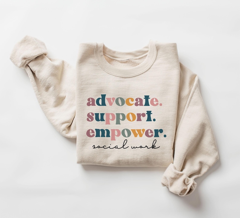 May include: Cream-colored sweatshirt with the words "advocate. support. empower. social work" in colorful text. The long sleeves are rolled up. The sweatshirt is made of a soft material.