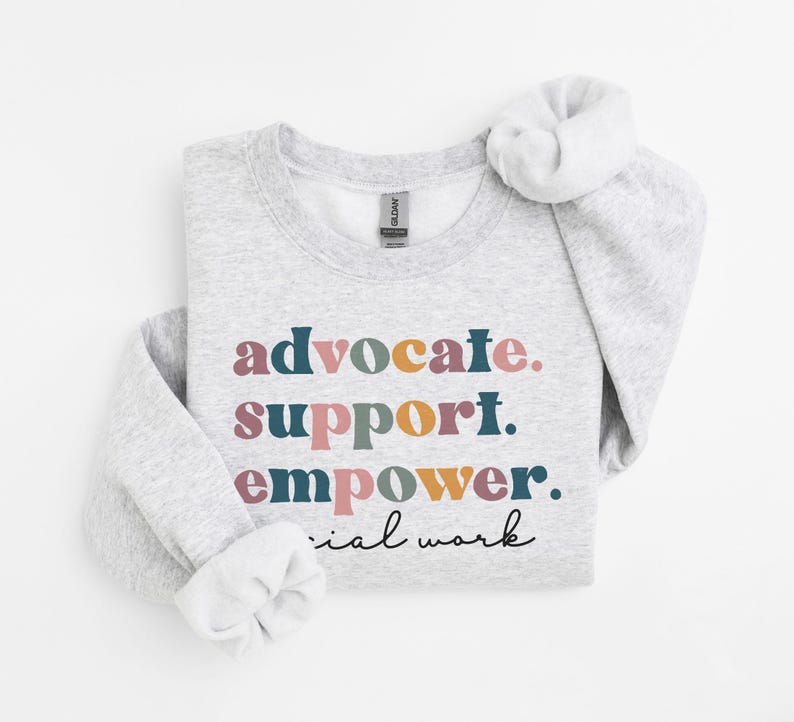 May include: A light gray sweatshirt with the words "advocate. support. empower. social work" in colorful text. The sweatshirt has a crew neck and long sleeves with rolled cuffs. The Gildan brand tag is visible.