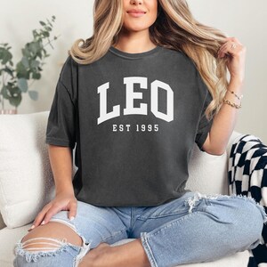 May include: Dark gray t-shirt with the word "LEO" in white block letters above "EST 1995". The shirt is worn with light blue jeans. The person is sitting on a white couch with a black and white checkered blanket.