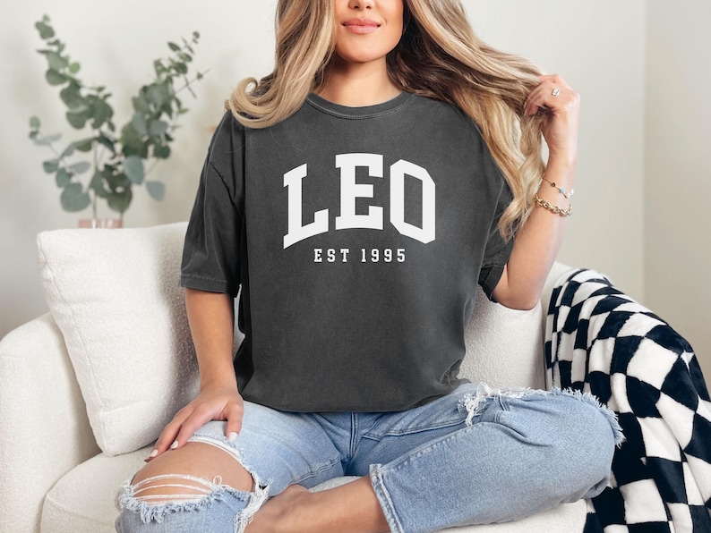 May include: Dark gray t-shirt with the word "LEO" in white block letters above "EST 1995". The shirt is worn with light blue jeans. The person is sitting on a white couch with a black and white checkered blanket.