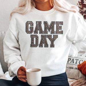 Pu&ograve; includere: Felpa bianca con il testo "GAME DAY" in lettere nere bordate con un motivo leopardato all'interno delle lettere.