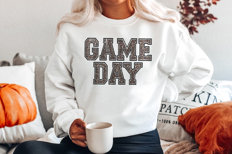 Pu&ograve; includere: Felpa bianca con il testo "GAME DAY" in lettere nere bordate con un motivo leopardato all'interno delle lettere.