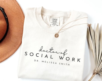 May include: A beige t-shirt with the text "doctor of SOCIAL WORK DR. MELISSA SMITH" printed on the front. The shirt is laid flat on a white surface.