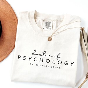 May include: A beige t-shirt with the text "doctor of PSYCHOLOGY" and "DR. MICHAEL JONES" printed in black.