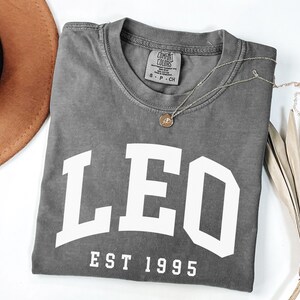 May include: A gray Comfort Colors t-shirt with the word "LEO" in large white block letters and "EST 1995" underneath. A gold necklace with a pendant rests on the shirt. A brown hat and dried palm leaf are in the background.