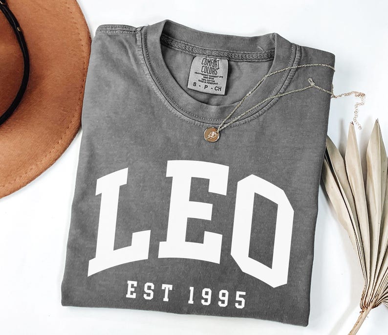 May include: A gray Comfort Colors t-shirt with the word "LEO" in large white block letters and "EST 1995" underneath. A gold necklace with a pendant rests on the shirt. A brown hat and dried palm leaf are in the background.