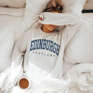 以下が含まれることがあります： 「EDINBURGH」の文字がネイビーブルーのブロック体で、その下に「SCOTLAND」と書かれたライトグレーのスウェットシャツ。白い寝具のベッドに横たわり、コーヒーカップを持っています。顔は腕で覆われています。
