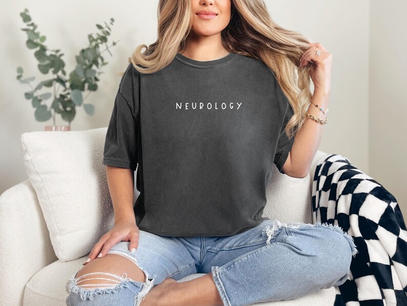 K&ouml;nnte beinhalten: Ein dunkelgraues, &uuml;bergro&szlig;es T-Shirt mit dem Wort "NEUROLOGY" in wei&szlig; auf der Vorderseite. Das Shirt wird von einer Person getragen, die auf einem wei&szlig;en Sofa mit einer schwarz-wei&szlig;en karierten Wolldecke sitzt.