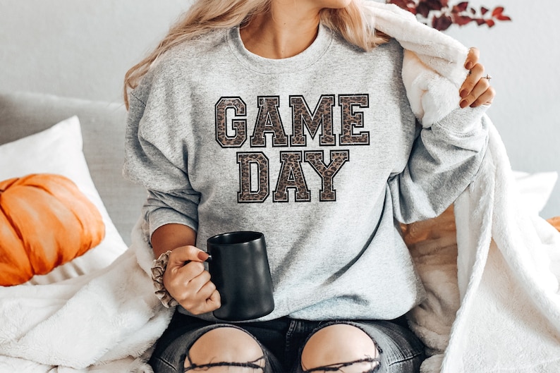 Pu&ograve; includere: Una felpa grigia con la scritta "GAME DAY" stampata in lettere nere con un motivo leopardato.