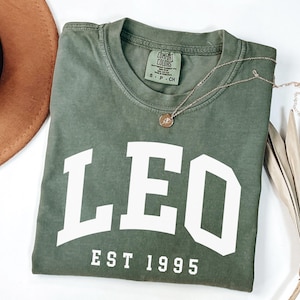 May include: Olive green t-shirt with the word "LEO" in white block letters and "EST 1995" underneath. A gold necklace with a pendant rests on the shirt. A brown hat and dried palm leaf are in the background.