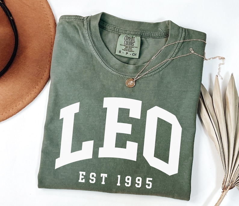 May include: Olive green t-shirt with the word "LEO" in white block letters and "EST 1995" underneath. A gold necklace with a pendant rests on the shirt. A brown hat and dried palm leaf are in the background.