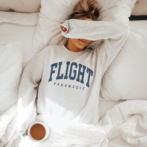 May include: A light gray sweatshirt with the words "FLIGHT PARAMEDIC" in navy blue block letters. The sweatshirt is worn on a person lying in bed with white bedding. A cup of coffee sits on the bed.