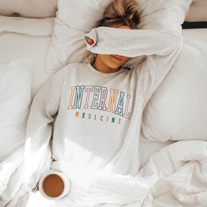 May include: A light gray sweatshirt with the words "INTERNAL MEDICINE" in colorful lettering. The person is lying on a bed with white bedding, with a cup of coffee nearby.