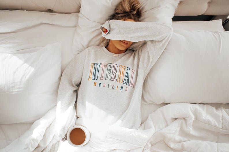Puede incluir: Una sudadera gris claro con las palabras "INTERNAL MEDICINE" en letras coloridas. La persona est&aacute; acostada en una cama con ropa de cama blanca, con una taza de caf&eacute; cerca.